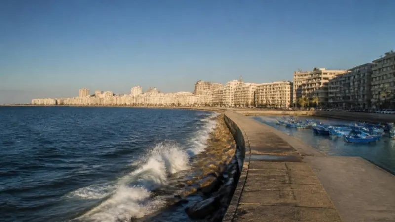 Waves breaking on the sea wall of the Alexandria Corniche, with apartment buildings curving along the Mediterranean bay